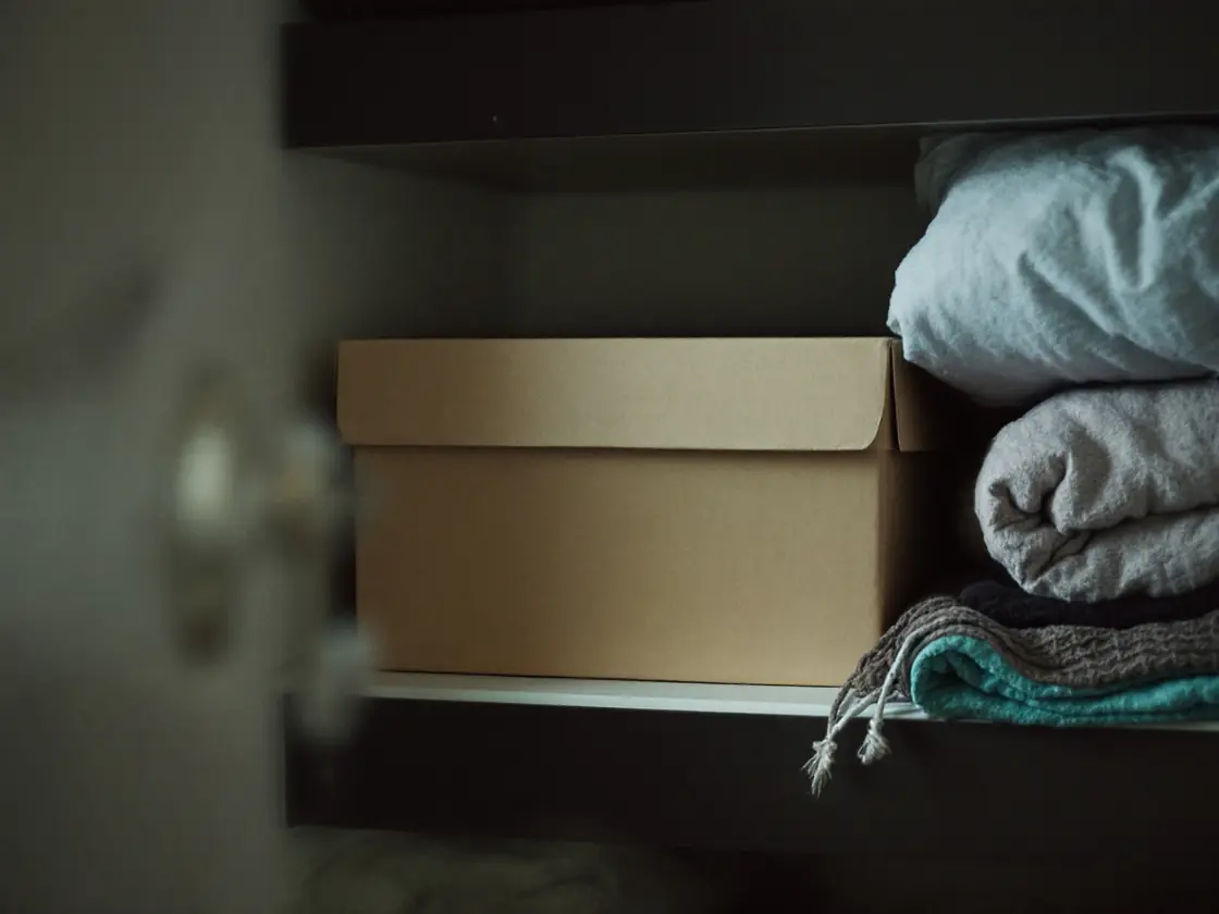 A closed plain shoebox on a closet shelf beside folded blankets